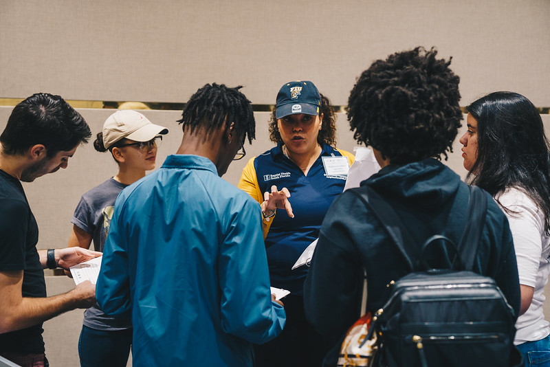 Group of students standing in a circle facing each other, all of them seem to be speaking over one another while one student, a female with curly hair wearing an FIU hat, points to the back of the class for everyone to turn and see.