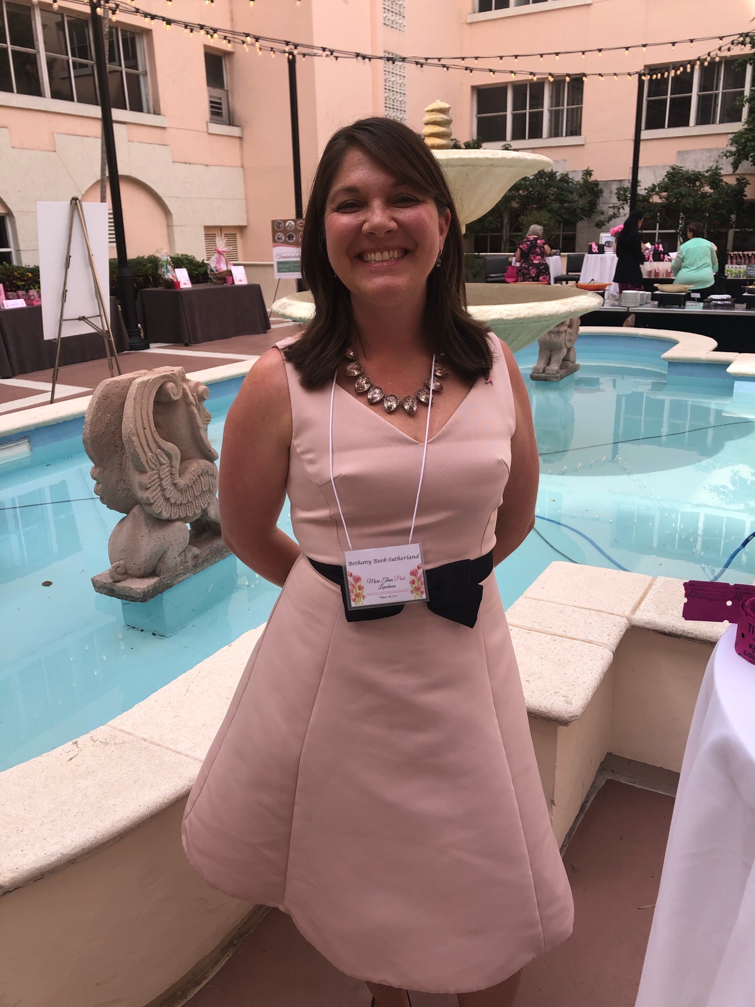 Dr. Bethany Sutherland standing in front of a large garden fountain, she is wearing a long pink dress and has her hands behind her back. She is smiling widely and standing with confidence. 