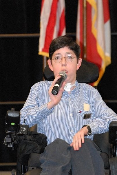 zach trantenberg sitting in his wheelchair in front of a group with a microphone in his hand