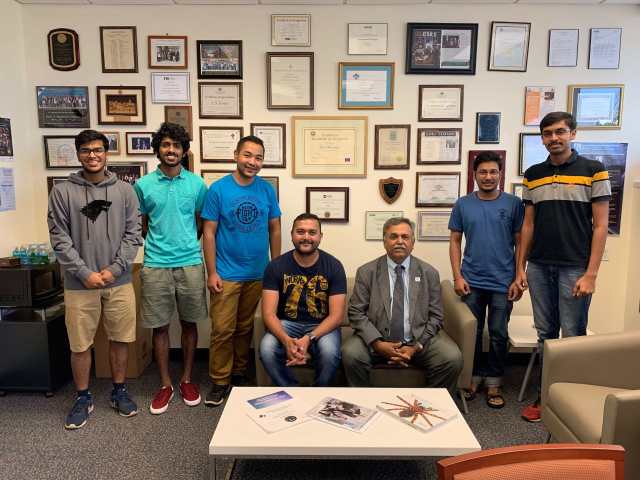 Six students and one professor from the Science without Borders Summer Program are posing in front of a well decorated with plaques and awards