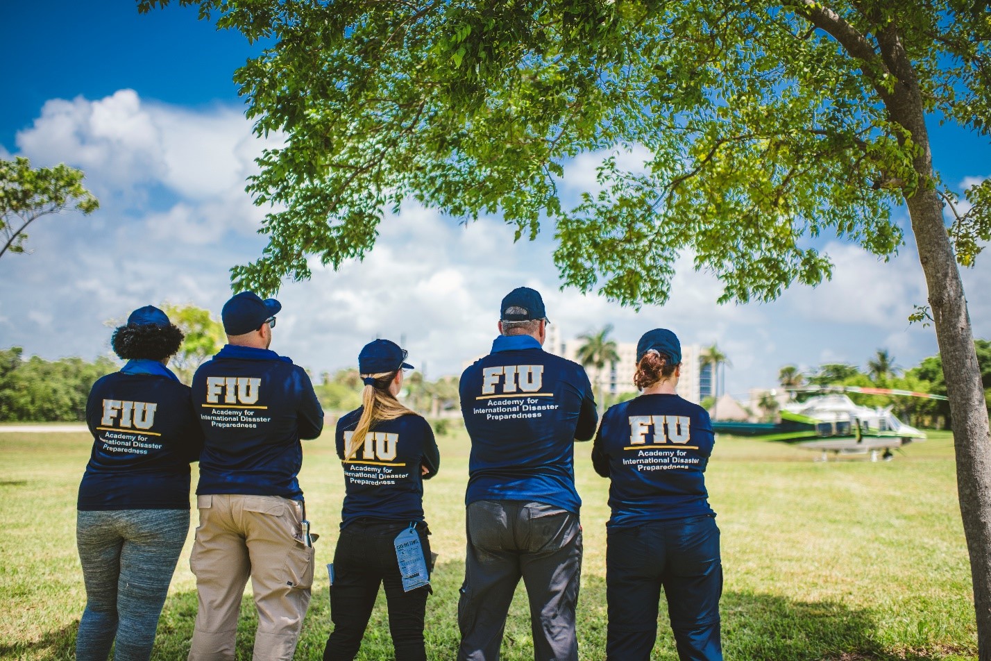 A group of 8 students standing in a medium length grassy field looking towards the sky with their backs to the camera all of them wearing navy blue polos that ready Academy of Disaster Management  