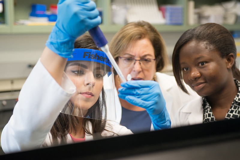 Dr. Huffman standing in between two female students in lab coats and with protective eye gear as they use a drip to suction up a sample for testing